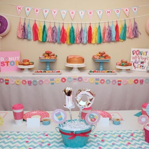 May include: A donut-themed birthday party table setting with pink and blue decorations. The table is decorated with a donut garland, donut balloons, and a donut-shaped cake. There are also two pink and blue buckets with white tissue paper and a silver number 10 balloon. The table is set with pink and blue plates, napkins, and cups. The table is covered with a white tablecloth with a pink and blue chevron pattern.