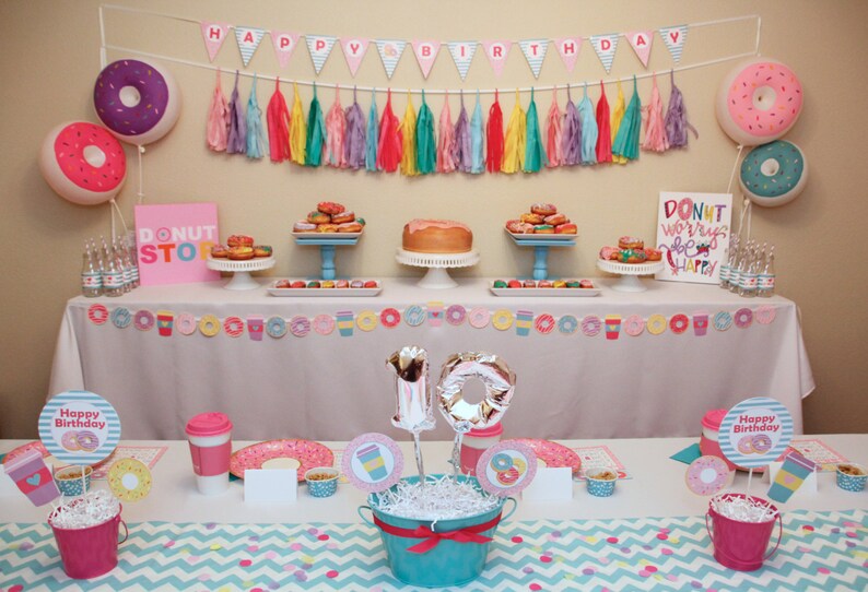 May include: A donut-themed birthday party table setting with pink and blue decorations. The table is decorated with a donut garland, donut balloons, and a donut-shaped cake. There are also two pink and blue buckets with white tissue paper and a silver number 10 balloon. The table is set with pink and blue plates, napkins, and cups. The table is covered with a white tablecloth with a pink and blue chevron pattern.