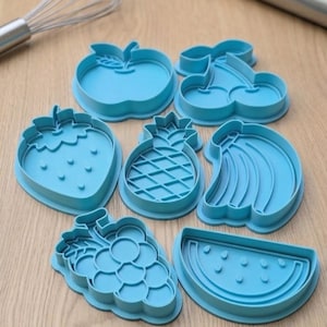May include: A set of seven light blue plastic fruit-shaped cookie cutters, including designs of an apple, cherries, a strawberry, a pineapple, a banana bunch, grapes, and a watermelon slice, arranged on a light wood surface with a metal whisk.
