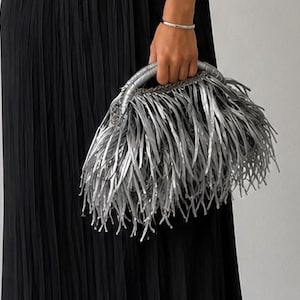 May include: A silver handbag with a fringe design is held by a person. The bag has a woven handle and is made of a metallic material. The fringe is long and flowing, creating a textured look. The person is wearing a black dress.