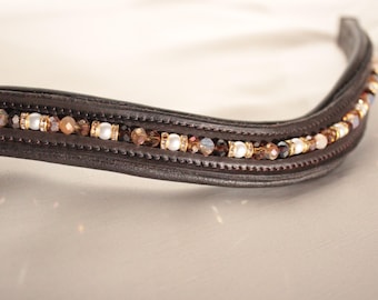 Könnte beinhalten: Ein braunes Leder-Stirnband für Pferde mit einer dekorativen Reihe aus weißen, goldenen und braunen Perlen.