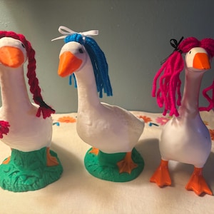 May include: Three whimsical goose figurines with white bodies, orange beaks and feet, and green bases. Each goose has yarn hair in red, blue, and pink. The geese are displayed on a floral patterned tablecloth.