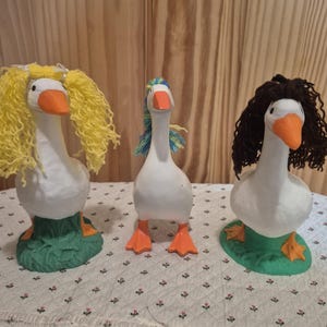 May include: Three whimsical goose figurines with white bodies, orange beaks and feet, and green bases. Each goose has a different hairstyle: yellow curly hair, blue and yellow hair, and brown curly hair. The figurines are on a white patterned surface.