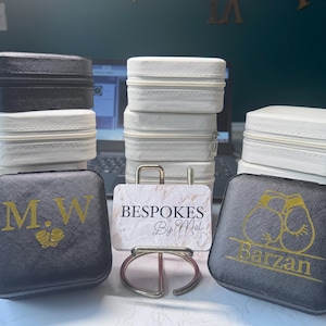 Può includere: Un assortimento di piccole custodie quadrate per gioielli in bianco e nero, impilate e disposte su una superficie. Alcune custodie presentano scritte e disegni dorati. Un porta biglietti da visita con una carta effetto marmo è al centro, con il testo "BESPOKES By Mel".