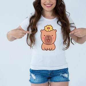 May include: White t-shirt featuring a cartoon bear with a flower on its head. The bear is brown with a pink nose and tongue. The shirt is paired with denim shorts.