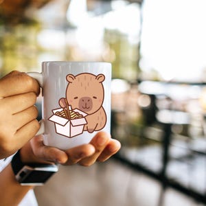 May include: A white ceramic mug featuring a cartoon bear illustration. The bear is brown and holding chopsticks and a box of noodles. The mug is held by a person.