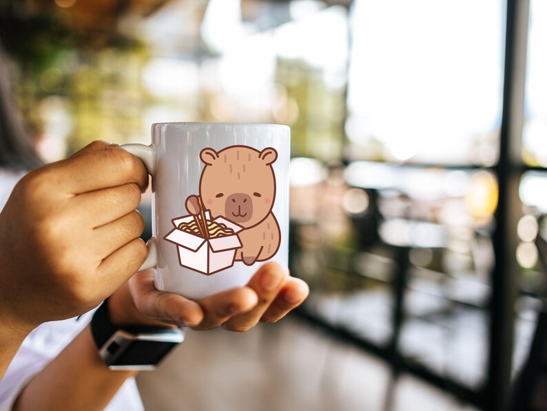 May include: A white ceramic mug featuring a cartoon bear illustration. The bear is brown and holding chopsticks and a box of noodles. The mug is held by a person.