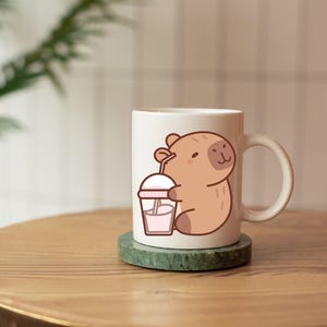 May include: A white ceramic mug featuring a cartoon capybara holding a pink drink with a straw. The mug sits on a green coaster and is on a wooden surface. The capybara is light brown with a smiling face.