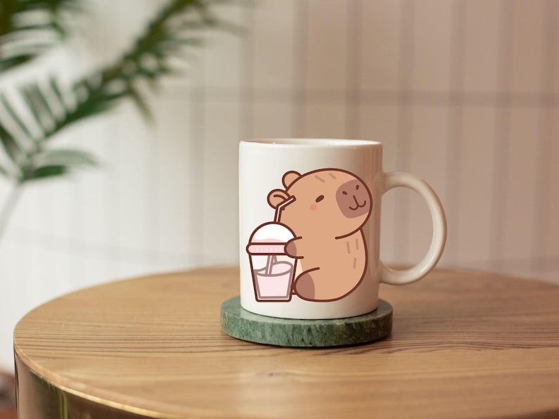 May include: A white ceramic mug featuring a cartoon capybara holding a pink drink with a straw. The mug sits on a green coaster and is on a wooden surface. The capybara is light brown with a smiling face.