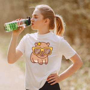 May include: White t-shirt featuring a cartoon capybara wearing yellow sunglasses and a star. The capybara design is centered on the shirt. A clear water bottle with green accents is being held up to the mouth.