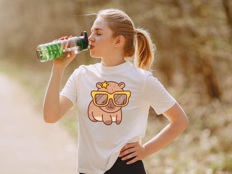 May include: White t-shirt featuring a cartoon capybara wearing yellow sunglasses and a star. The capybara design is centered on the shirt. A clear water bottle with green accents is being held up to the mouth.