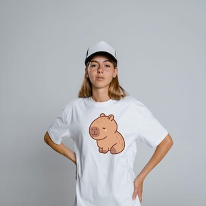 May include: White t-shirt featuring a cartoon capybara illustration in shades of brown. The capybara is sitting with a smiling expression. The person in the image is wearing a white and black trucker hat.