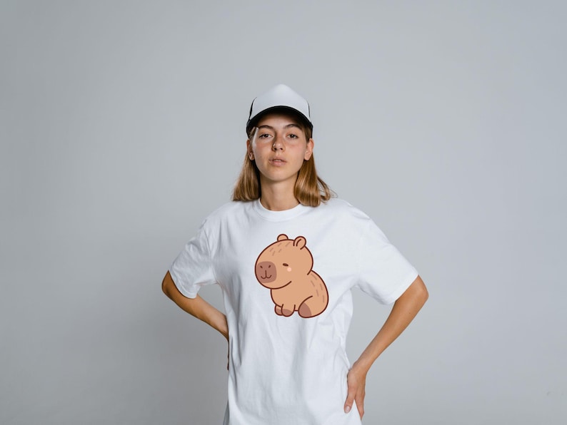 May include: White t-shirt featuring a cartoon capybara illustration in shades of brown. The capybara is sitting with a smiling expression. The person in the image is wearing a white and black trucker hat.