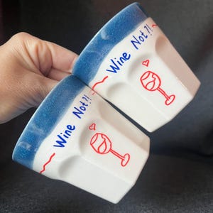 May include: Two white ceramic wine glasses with a blue rim. Each glass has the words "Wine Not?!" in blue, a red line, a red wine glass graphic, and a heart.