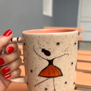 May include: A ceramic mug with a speckled cream-colored exterior and a solid peach-colored interior. The mug features a hand-painted black and orange figure of a person dancing, accented by small raised black dots across the surface.