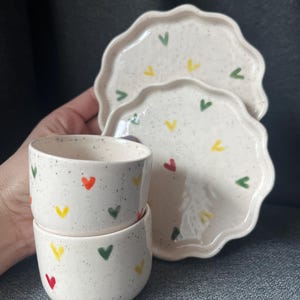 May include: A set of handmade ceramic tableware featuring two small cups and two scalloped-edge plates. The off-white pieces are speckled with dark flecks and decorated with colorful heart-shaped designs in red, yellow, and green. The set is designed for serving small portions.