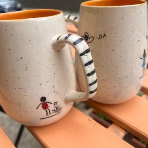 Puede incluir: Dos tazas de cerámica con exteriores beige moteado e interiores naranjas. Una taza tiene un asa a rayas blancas y negras, y ambas tienen ilustraciones caprichosas de personas y notas musicales. Las tazas están sobre una superficie naranja.