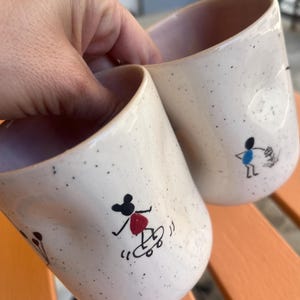 May include: Two speckled ceramic mugs with a light pink interior. Each mug features a black and red cartoon figure skateboarding. The mugs are held by a person's hand.