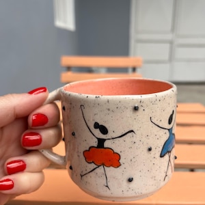Puede incluir: Una taza de cerámica con exterior crema, salpicada de puntos negros e interior melocotón. La taza presenta ilustraciones dibujadas a mano de bailarinas con tutús naranja y azul. La taza es sostenida por una persona con esmalte de uñas rojo.