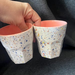 May include: Two white ceramic cups with a speckled design of blue, yellow, and orange dots. The interior of each cup is a solid coral color. The cups have a geometric, faceted shape.