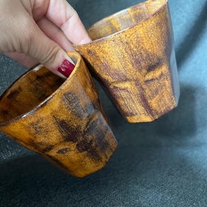May include: Two handmade ceramic cups with a textured, mottled finish in shades of brown and gold. The cups have a unique, angular design and are held by a hand with red nail polish. These cups are perfect for coffee, tea, or other beverages.