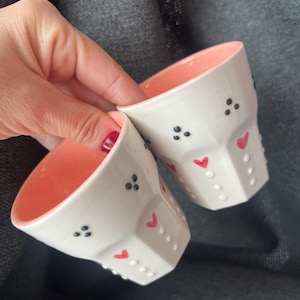 May include: Two white ceramic cups with a pink interior. The cups have a geometric design with black dots and red heart accents. The cups are held against a gray background.
