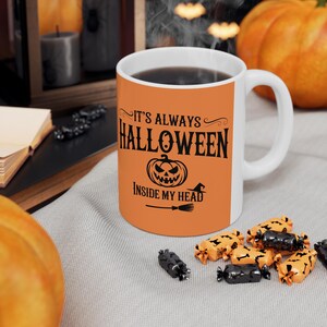 May include: White ceramic mug with an orange band featuring the text "IT'S ALWAYS HALLOWEEN INSIDE MY HEAD" in black, along with a jack-o'-lantern and witch's broom. The mug is filled with a dark liquid and surrounded by Halloween candy.