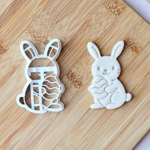 Puede incluir: Dos cortadores de galletas blancos con forma de conejito sobre una tabla de cortar de madera. Un cortador es un diseño de conejito sólido, y el otro es un conejito con un huevo estampado. Los cortadores son para hornear y golosinas temáticas de Pascua.