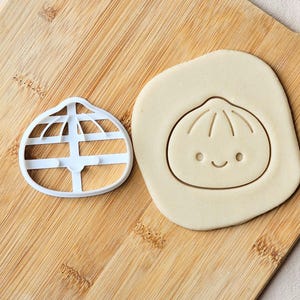 May include: A white plastic dumpling-shaped cookie cutter next to a piece of rolled-out dough that has been imprinted with a cute, smiling dumpling face design. The cutter features a rounded top with a flat base and internal structural supports.