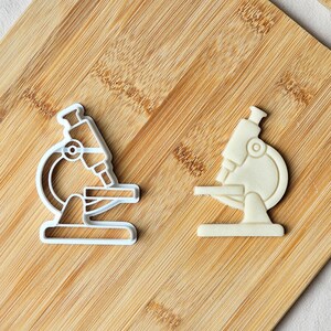 May include: Two microscope-shaped cookie cutters. One is white plastic, the other is a white outline. The cutters are on a light brown wooden cutting board. The microscope design includes the eyepiece, objective lenses, and base.