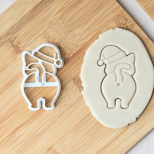 May include: A white plastic cookie cutter in the shape of a Santa Claus-themed cat, with a matching cutout in a piece of dough. The cutter is on a wooden cutting board.