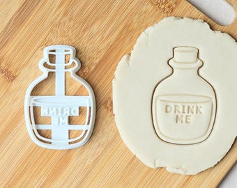 May include: A white plastic bottle-shaped cookie cutter next to a piece of dough embossed with the same bottle shape and the text DRINK ME. The cutter features a rounded base, a narrow neck, and a stopper top, creating a clear outline and imprint.