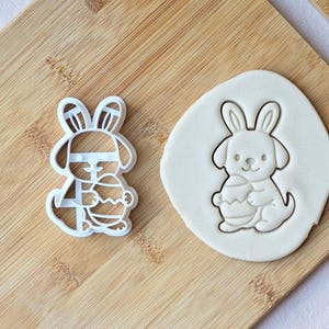 May include: A white plastic cookie cutter shaped like a bunny dog holding an Easter egg, alongside a cookie cut-out on a wooden board. The cutter is about 7.6 cm tall.