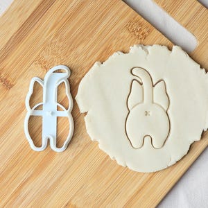 May include: A light blue plastic cookie cutter shaped like a cat's rear end is next to a piece of dough with the same shape. The cutter is on a wooden chopping board.