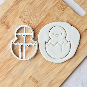 May include: A white plastic chick-in-egg cookie cutter next to a chick-shaped imprint in dough. The cutter is a detailed outline of a chick hatching from an egg. The dough imprint shows the chick's features.