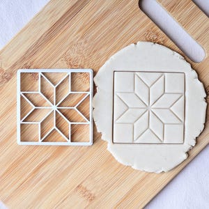 May include: A white square cookie cutter featuring an eight-pointed star geometric pattern, positioned next to a piece of rolled-out white dough that has been imprinted with the same star design, both resting on a light brown wooden cutting board.