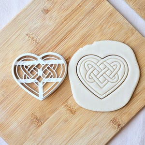 May include: A white heart-shaped cookie cutter featuring an intricate Celtic knot design inside the frame, positioned next to a piece of white dough that has been stamped with the same heart-shaped Celtic knot pattern to show the impression.