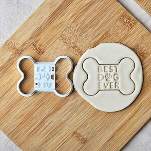 以下が含まれることがあります： 中央に肉球のアイコンと「BEST DOG EVER」の文字が入った、骨の形をした白いプラスチック製のクッキー型。その横には、同じ骨の形と文字が刻印された円形の明るい色の生地が置かれています。
