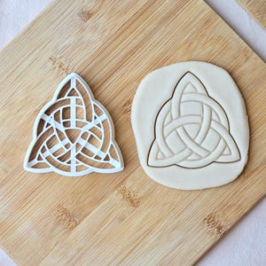 Könnte beinhalten: Ein weißer Kunststoff-Ausstecher mit einem dreieckigen keltischen Triquetra-Knotenmuster, platziert neben einem kreisförmigen Stück ausgerolltem Teig, das mit demselben komplexen, ineinander verschlungenen Knotenmuster geprägt ist.