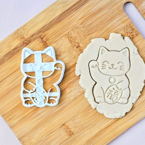 May include: A light blue plastic cookie cutter shaped like a waving cat, alongside a cookie with the same design imprinted. The cutter and cookie are on a wooden chopping board.