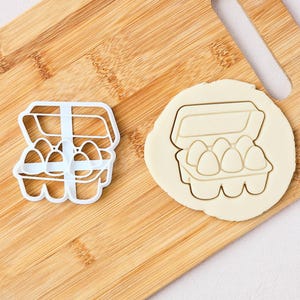 May include: A white plastic cookie cutter shaped like an open egg carton containing six eggs, alongside a circular piece of rolled-out dough that has been stamped with the same egg carton design, showing the clear outline of the container and eggs.