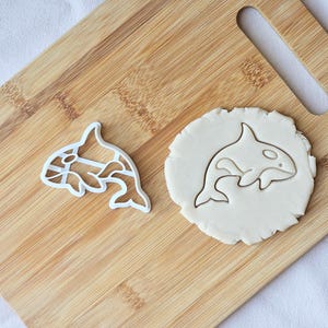 May include: A white plastic orca cookie cutter and a round piece of dough with an orca imprint. The cutter has a detailed design, while the dough shows a simple outline. The items are on a wooden cutting board.