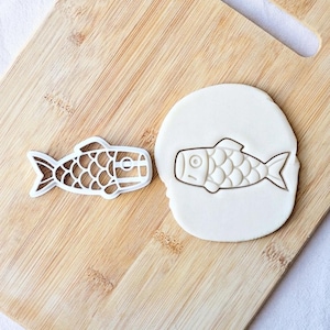 May include: A white plastic fish-shaped cookie cutter next to a circular piece of white dough featuring an embossed fish design. The fish shape has a detailed scale pattern, a distinct eye, and a tail fin, creating a clear impression in the dough.