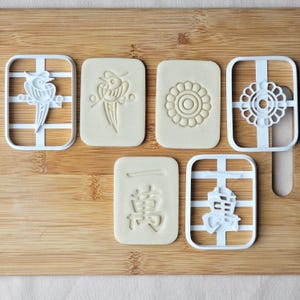 K&ouml;nnte beinhalten: Sechs Ausstecher und Kekse sind auf einer Holzoberfl&auml;che angeordnet. Die Ausstecher sind aus wei&szlig;em Kunststoff und die Kekse sind beige. Die Kekse haben gepr&auml;gte Designs eines Vogels, einer Blume und chinesischer Schriftzeichen. Die Ausstecher haben verschiedene Formen.