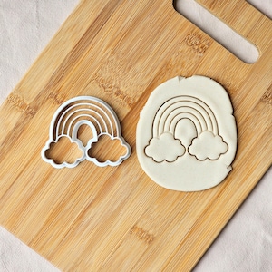 May include: A white plastic rainbow cookie cutter with a cloud at each end, positioned next to a piece of white dough that has been pressed with the same rainbow and cloud shape, creating a clear embossed outline on the flat surface.