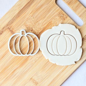 May include: A white pumpkin-shaped cookie cutter and a pumpkin-shaped imprint in dough are on a wooden chopping board. The cutter is a wireframe, and the dough imprint shows the pumpkin's shape.