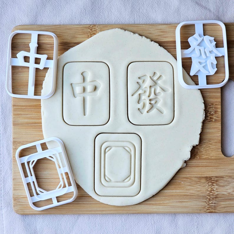 May include: A wooden cutting board with a sheet of dough and white plastic cookie cutters. The dough has impressions of Chinese characters and shapes. The cutters are in the shape of the characters and shapes.