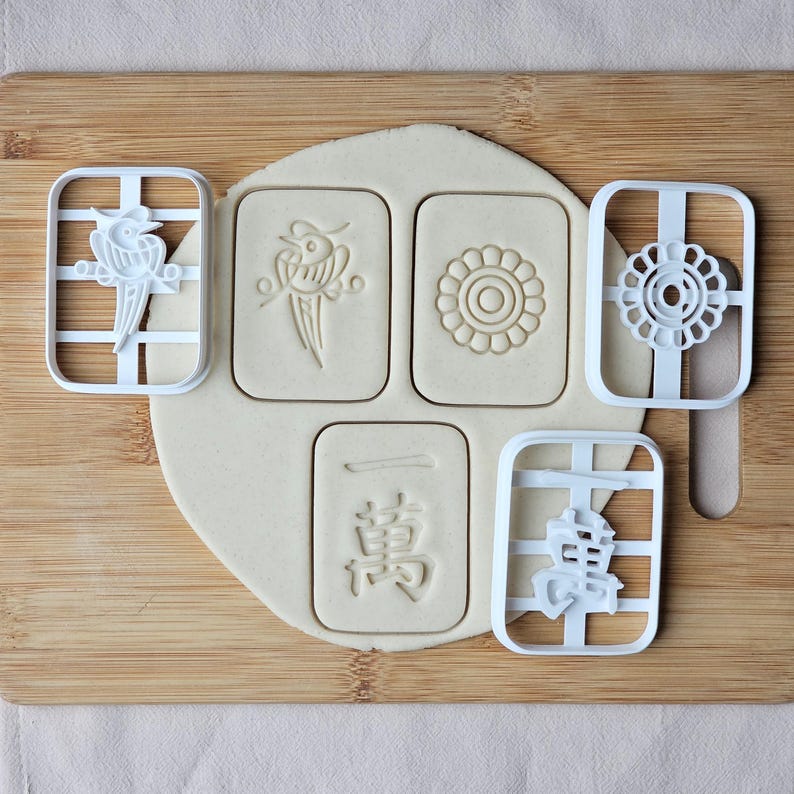 K&ouml;nnte beinhalten: Ein h&ouml;lzernes Schneidebrett mit einem Teigst&uuml;ck und f&uuml;nf wei&szlig;en Ausstechern. Die Ausstecher haben die Form von Mahjong-Steinen mit verschiedenen Designs und chinesischen Schriftzeichen. Der Teig hat Abdr&uuml;cke von den Ausstechern.