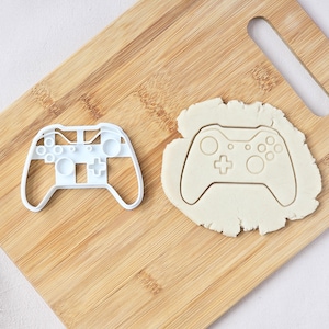 May include: A white plastic cookie cutter shaped like a video game controller is next to a controller-shaped cutout in dough. The cutter and dough are on a light brown wooden board.