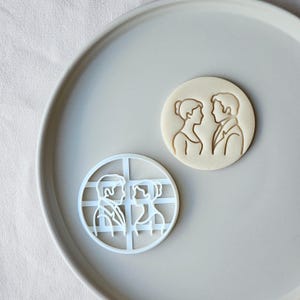 May include: A white plastic cookie cutter and a baked cookie with a couple design. The cookie cutter is round with a grid pattern. The baked cookie is round with a couple's silhouette.